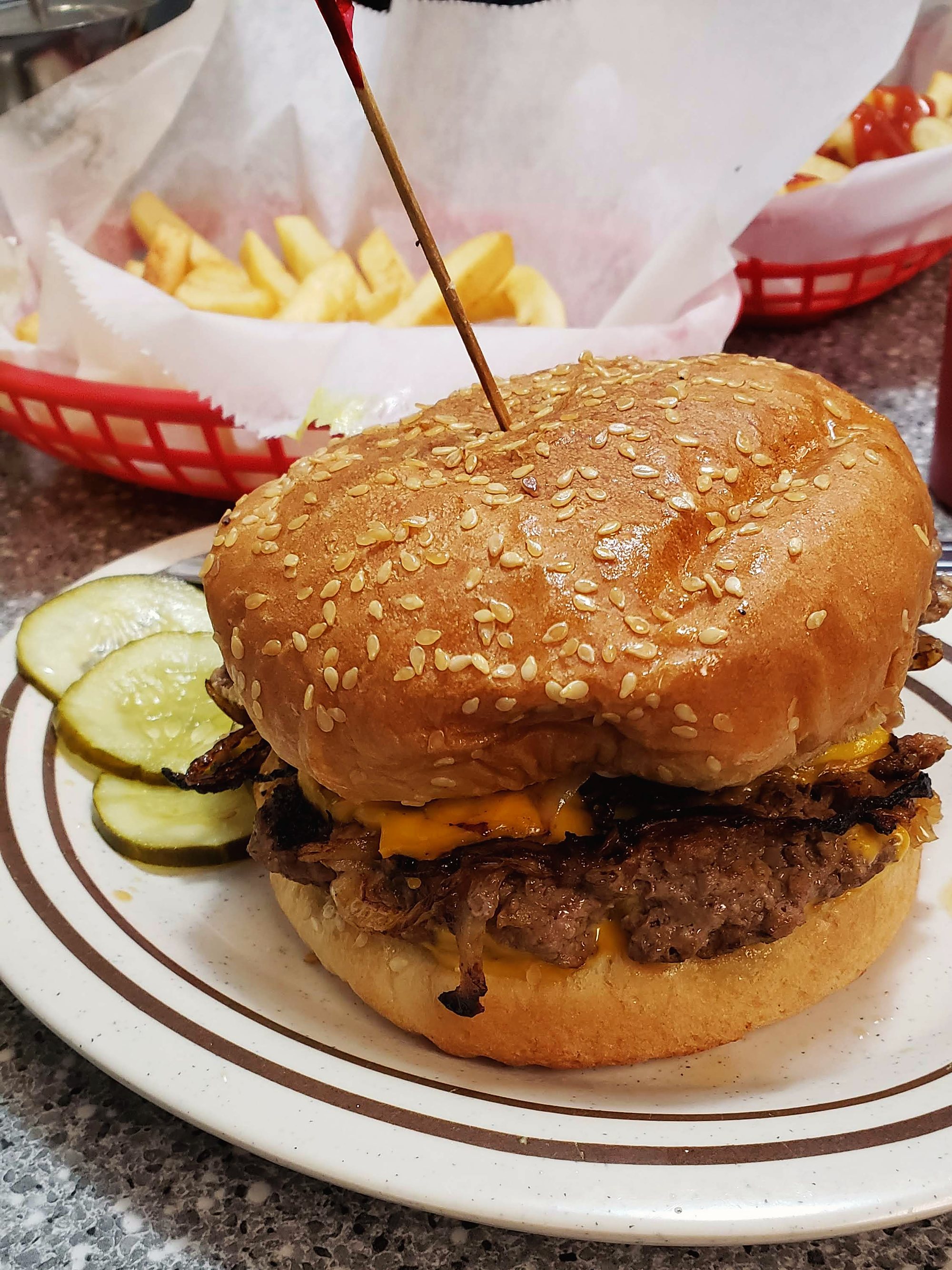 A Geneva's cheeseburger with pickles on a diner plate, a red basket of fries behind it.